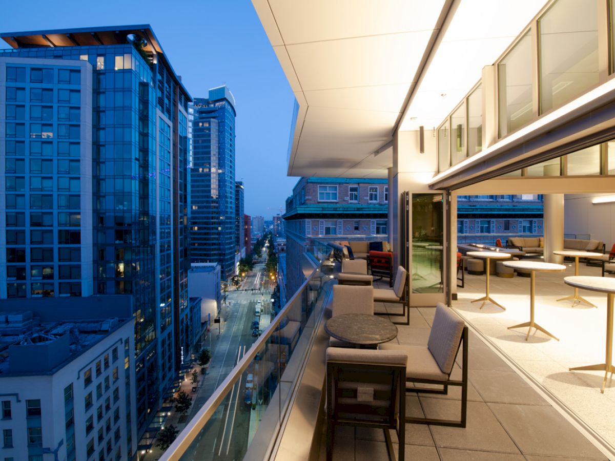 A modern cityscape at dusk viewed from a high-rise balcony with outdoor seating, tables, and surrounding tall buildings.
