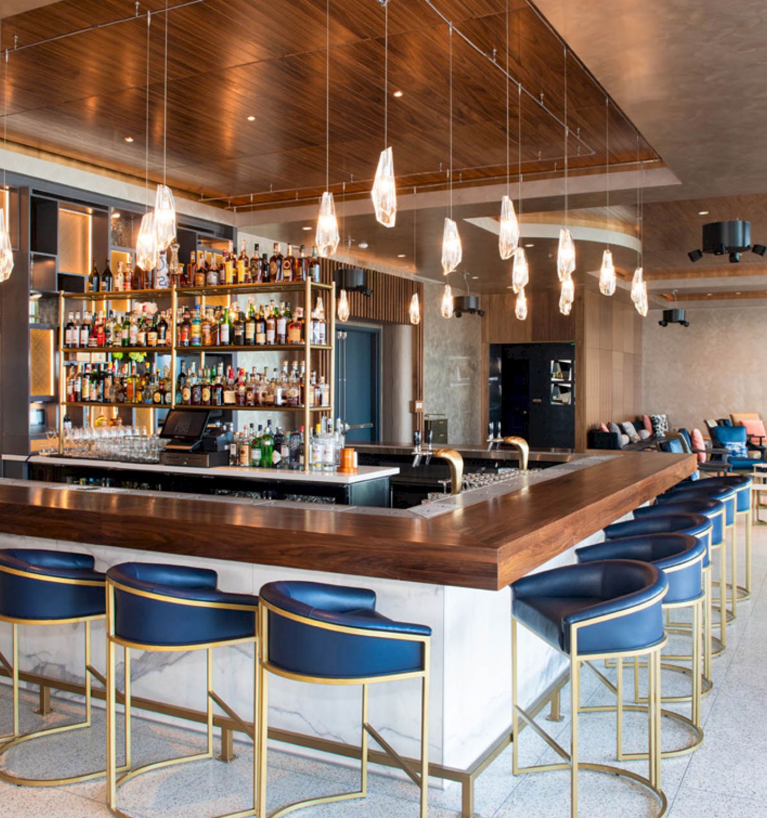This image shows a modern bar with a wooden countertop, blue bar stools, hanging lights, and a fully stocked liquor shelf in a stylish, open space.