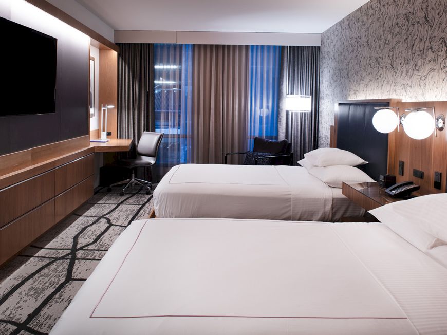The image shows a modern hotel room with two double beds, a flat-screen TV, a work desk, lamps, and floor-to-ceiling curtains.