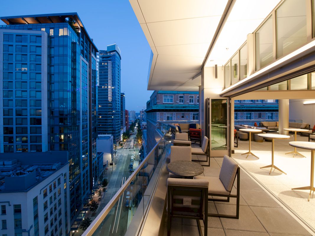 A modern urban balcony at twilight, featuring outdoor seating and tables, with a view of city buildings and a bustling street below.