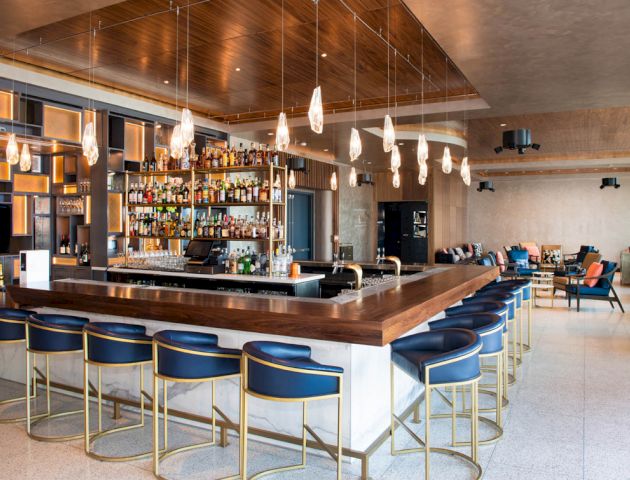 The image shows a modern bar with a stocked bar, stylish barstools, hanging lights, and a cozy seating area in the background.