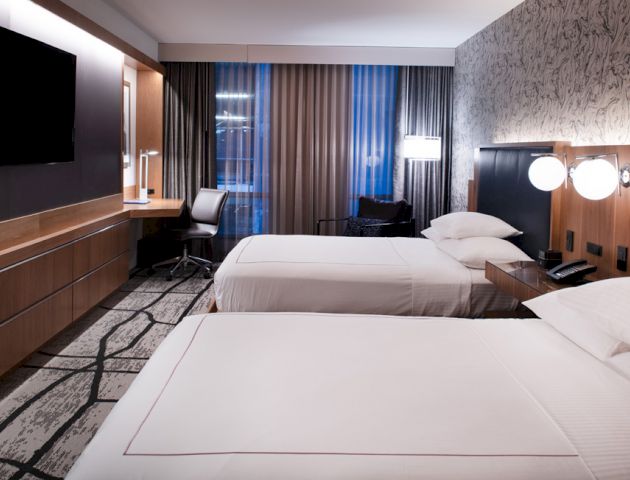 This image shows a modern hotel room with two neatly made beds, a desk, a TV on the wall, and contemporary decor.