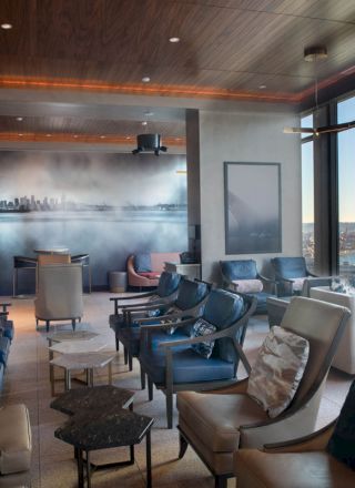 A modern, comfortable lounge with numerous chairs, tables, a wall mural, and large windows offering a scenic view outside during the daytime.