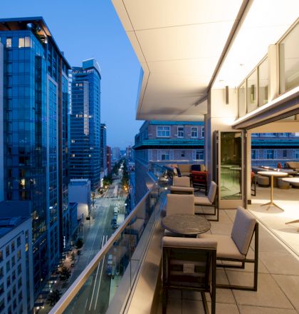 A modern balcony with seating and tables, overlooking a city street with tall buildings at dusk. The area looks inviting and well-lit.