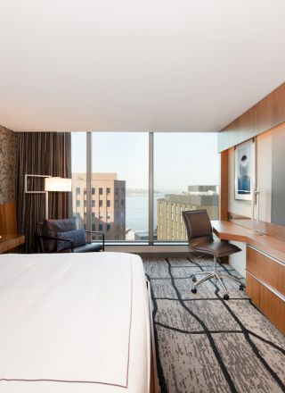 A modern hotel room with a large bed, wall-mounted TV, desk, chair, and window offering a city view with buildings and a glimpse of water.