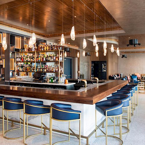 The image shows a modern bar with a wooden counter, blue chairs, hanging lights, and a well-stocked shelf of bottles behind the bar.