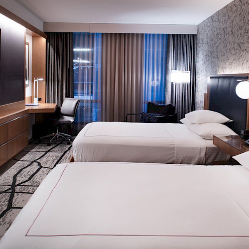The image shows a modern hotel room with two beds, a TV, a desk and chair, and a window with curtains. The room has stylish lighting fixtures.
