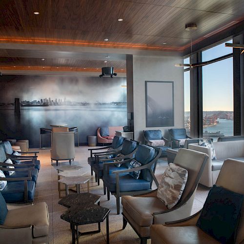 A modern lounge with multiple grey chairs, a cityscape mural, large windows offering a waterfront view, and warm wood accents.