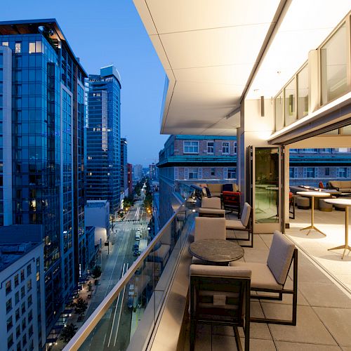 A modern urban balcony overlooks a cityscape with high-rise buildings, featuring outdoor seating and large windows leading into a lit interior.