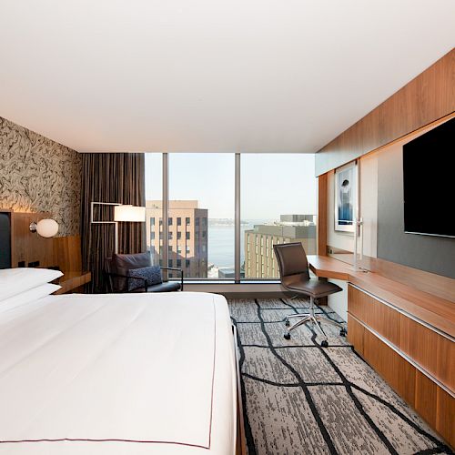 A modern hotel room featuring a large bed, desk, chair, flat-screen TV, and a window with a cityscape view, showcasing buildings and water.