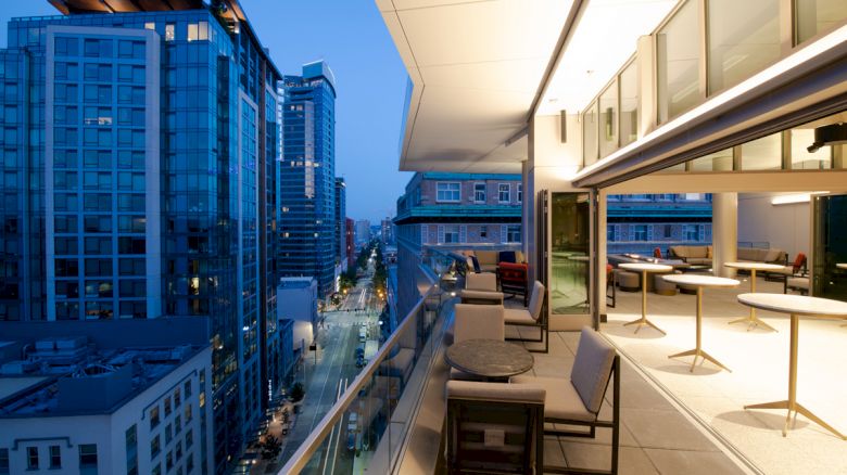 A modern balcony overlooking a city street with tall buildings, featuring chairs and tables, illuminated by evening lights.