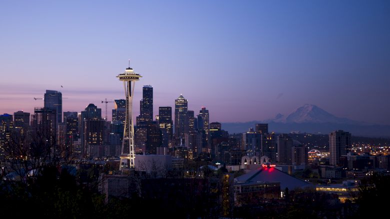 The image shows a twilight skyline of Seattle with the Space Needle and Mount Rainier in the background, featuring illuminated buildings.