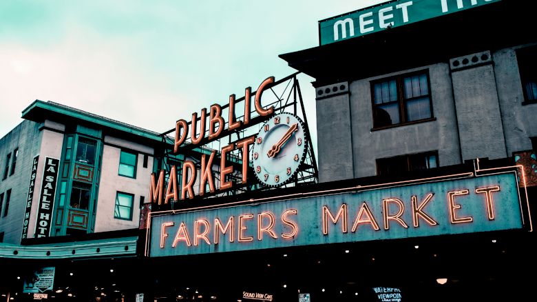 The image shows the neon signs for "Public Market" and "Farmers Market" at a well-known market area. The clock on the sign reads around 9:10.