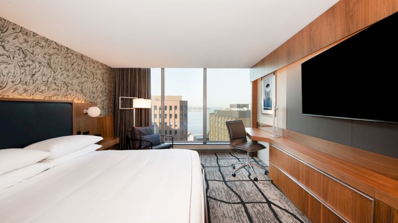 A modern hotel room with a large bed, wall-mounted TV, desk, chair, and window with city view. The walls have patterned decor.