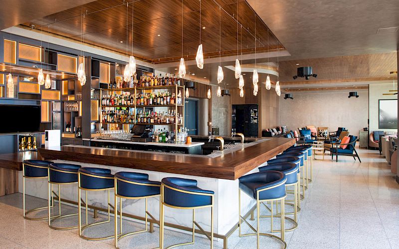 This image shows a modern bar with blue high chairs, warm lighting, and a stocked bar area. There are tables and seating areas in the background.