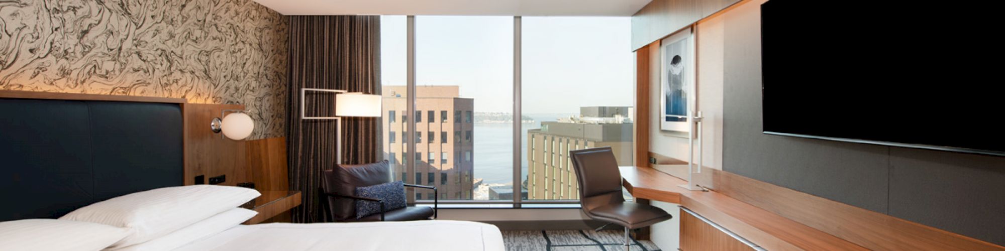 A modern hotel room with a large bed, a wall-mounted TV, a desk, a chair, and a window offering a city view.