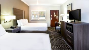 The image shows a hotel room with two beds, a TV, a desk, a chair, a window, and a door. The room has modern furnishings and carpeted flooring.