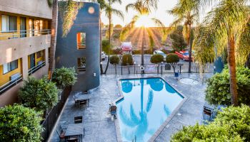 A serene outdoor pool area in a hotel with palm trees, surrounded by lounge chairs and tables, under a picturesque sunset.