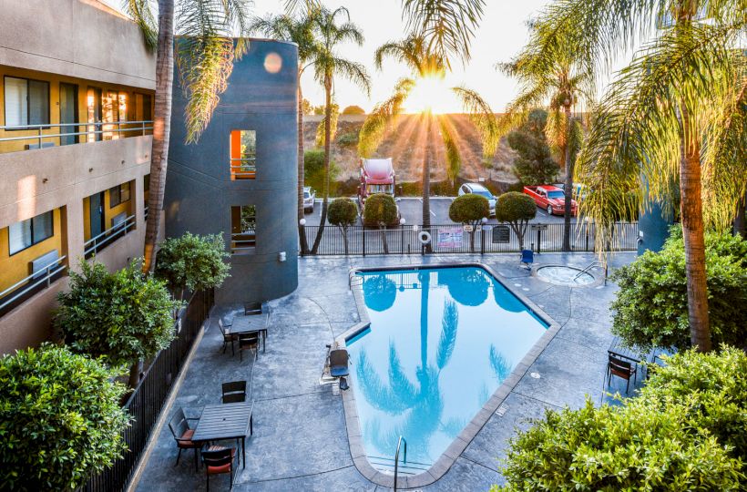 A serene outdoor pool area in a hotel with palm trees, surrounded by lounge chairs and tables, under a picturesque sunset.