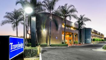 The image shows a Travelodge hotel building at dusk with palm trees and lighting. The hotel's sign is prominently displayed in the foreground.