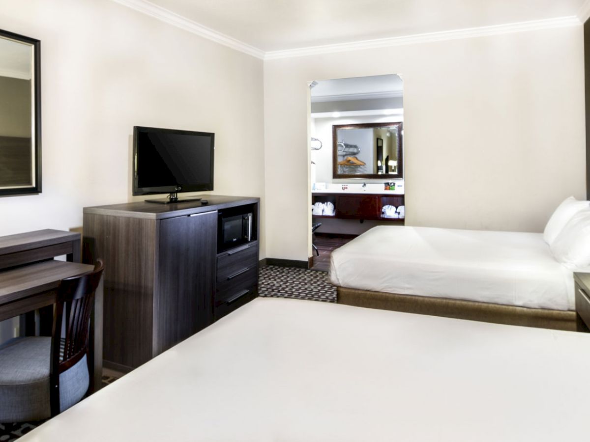 The image shows a modern hotel room with two beds, a desk, a TV on a console, and an open doorway leading to another room.