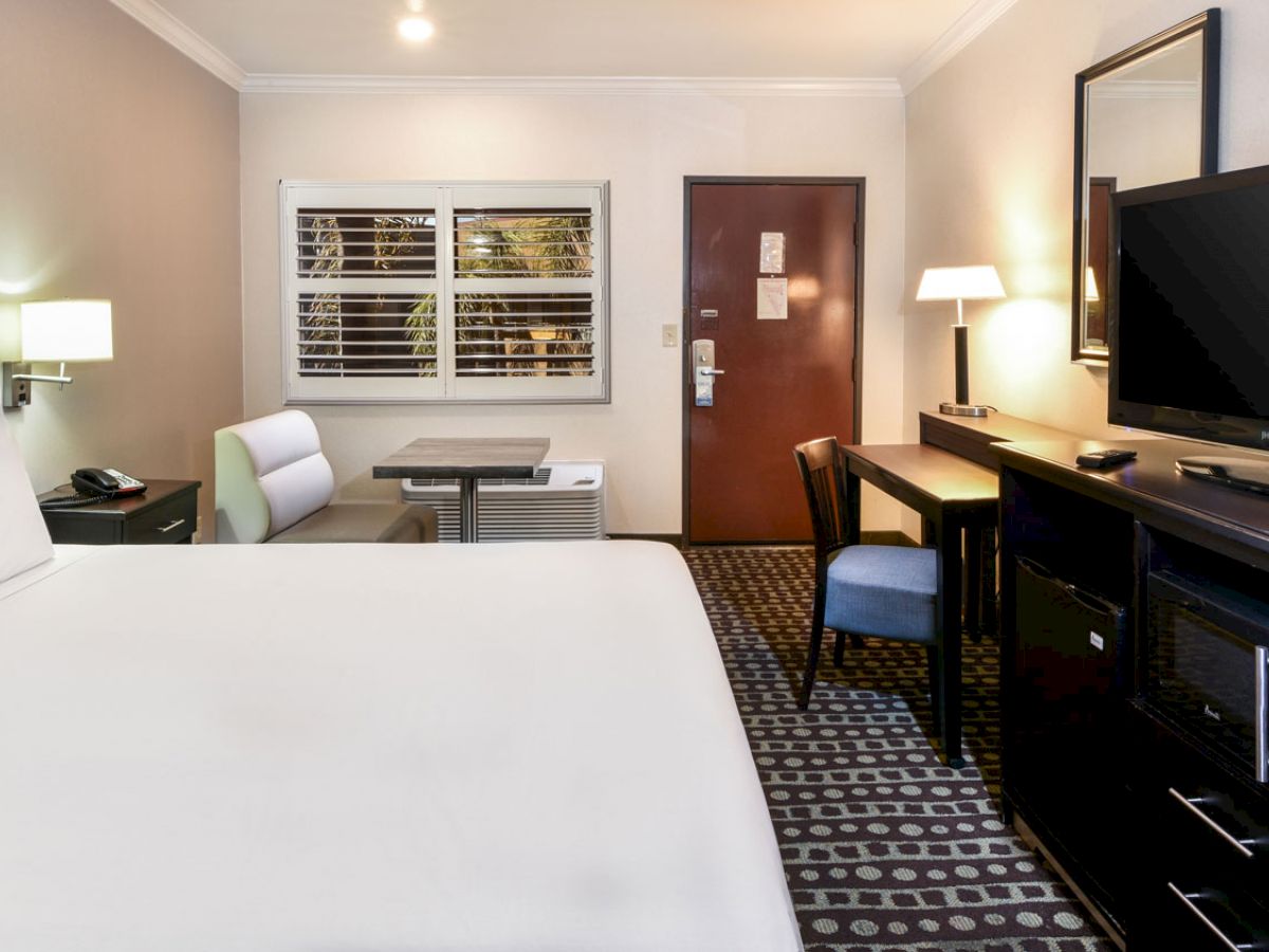 A well-furnished hotel room with a bed, desk, TV, and seating area near the door and window. Warm lighting enhances the cozy ambiance.