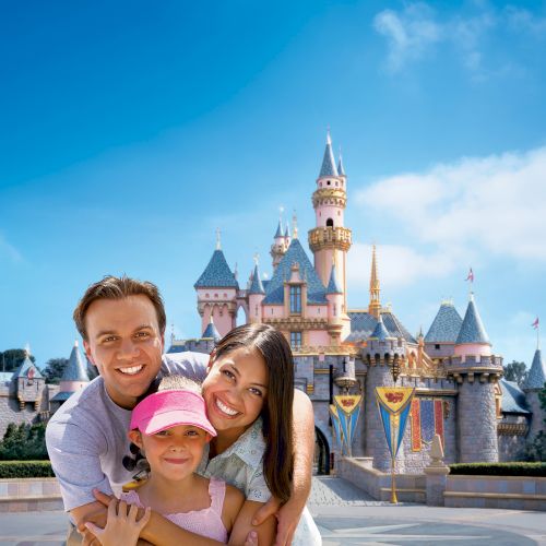 A smiling family posing in front of a castle-themed amusement park landmark on a clear day with a blue sky.