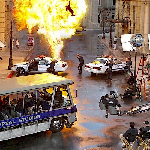 A movie set scene with an explosion, police cars, film crew, cameras, and a tram with people aboard at Universal Studios.