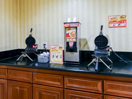 The image shows a breakfast area with waffle makers, a beverage dispenser, and cups on a countertop in front of a striped wall.