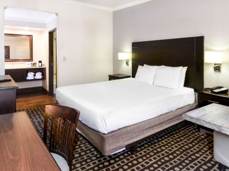 This image shows a hotel room with a king-sized bed, a desk with a chair, a flat-screen TV, bedside lamps, and a small seating area.