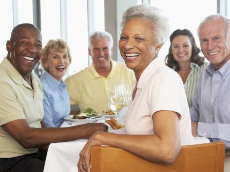 A group of smiling older adults sits around a table with food and drinks, appearing to enjoy a gathering in a well-lit, indoor setting.