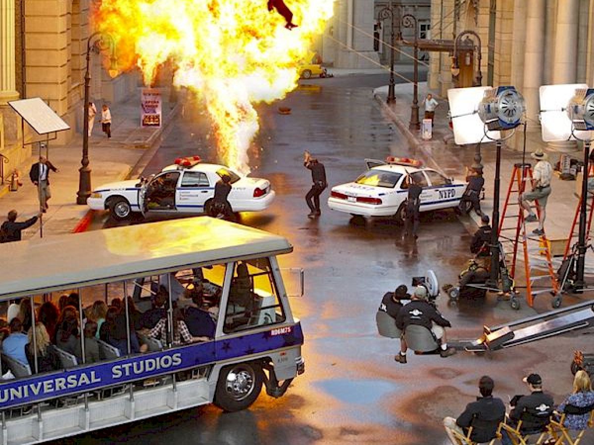 The image shows a movie set at Universal Studios with an explosion effect, police cars, actors, crew, and a tram full of tourists watching.