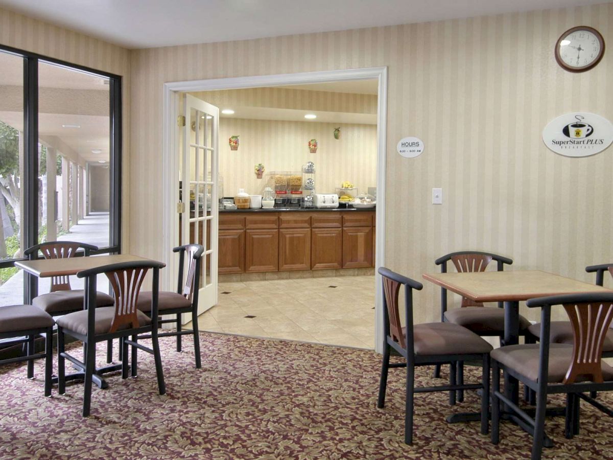 A small dining area with tables and chairs is adjacent to a room with a breakfast buffet; the space has a clock and wall décor.